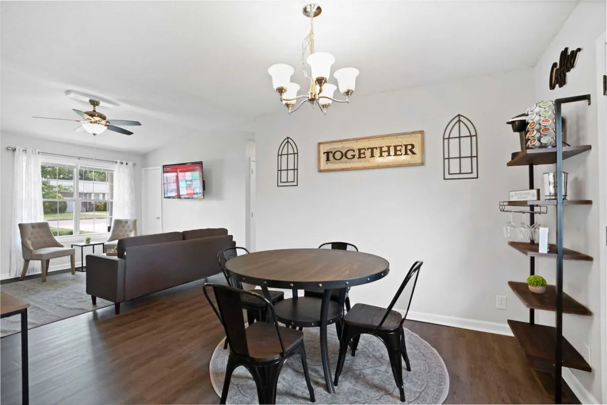 This image showcases a warmly lit dining table set beneath a chandelier, accented by a prominent 'TOGETHER' sign on the wall. A cozy sitting area with chairs and a TV is also visible, reinforcing the multi-purpose, inviting nature of the central living-dining hub.