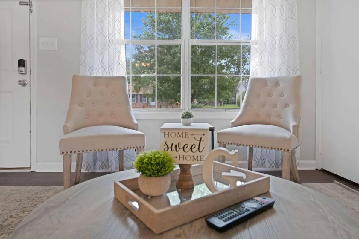 A cozy living area with two beige chairs flanking a wooden coffee table, topped with a decorative tray, plant, and a sign. Ample natural light streams in, creating an inviting and relaxed ambiance—ideal for unwinding after work or planning neighborhood outings.
