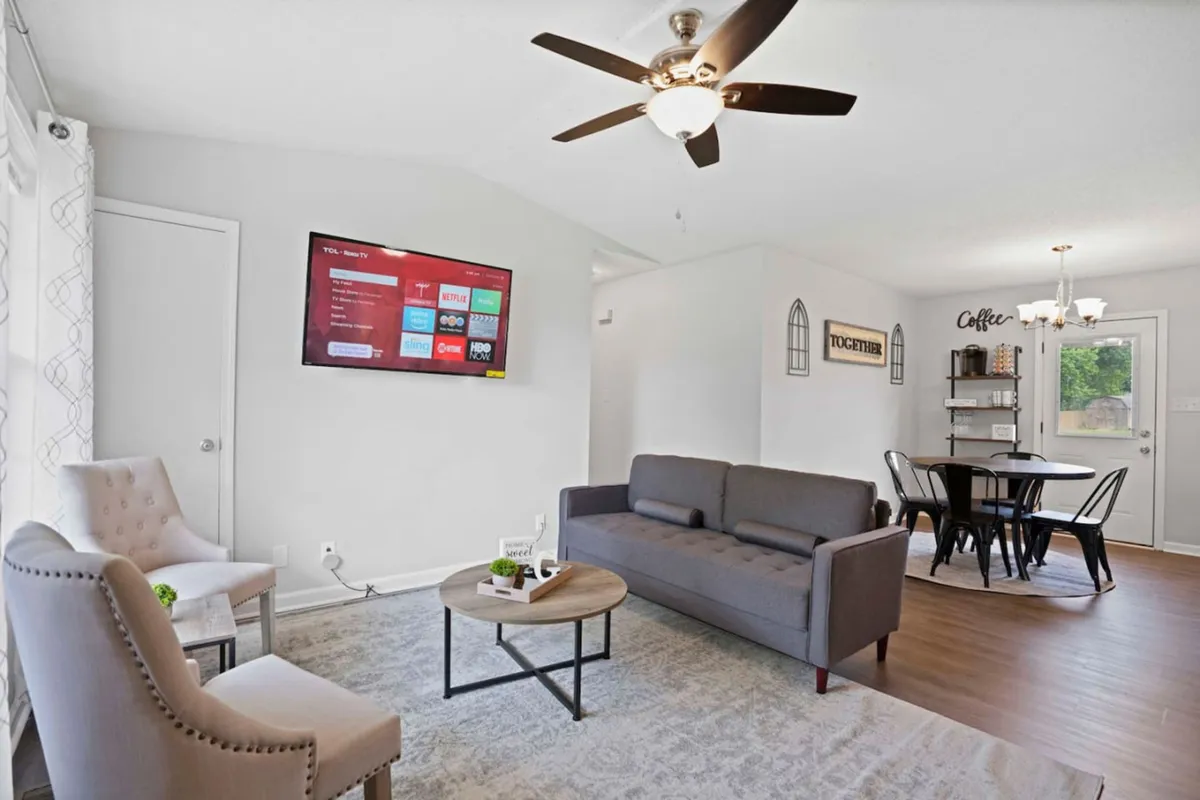 This image centers on a comfortable living area with a plush sofa, a coffee table, and a TV, while clearly showing the adjoining dining area in the background. Abundant natural light and tasteful décor create an inviting setting ideal for relaxation and socializing.