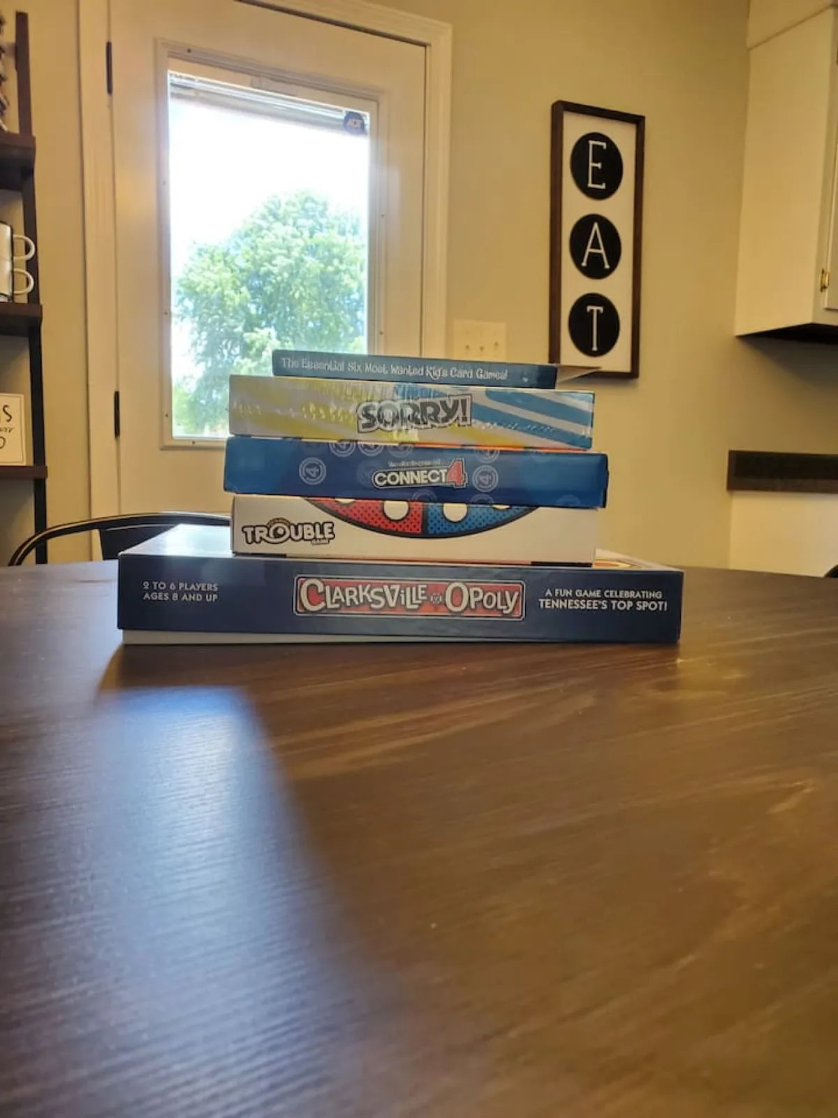 The image features a stack of board games on a table near a window, with sunlight streaming in and playful 'EAT' décor on the wall. The setup suggests lively family game nights and casual gatherings, reinforcing the room's multi-use appeal.
