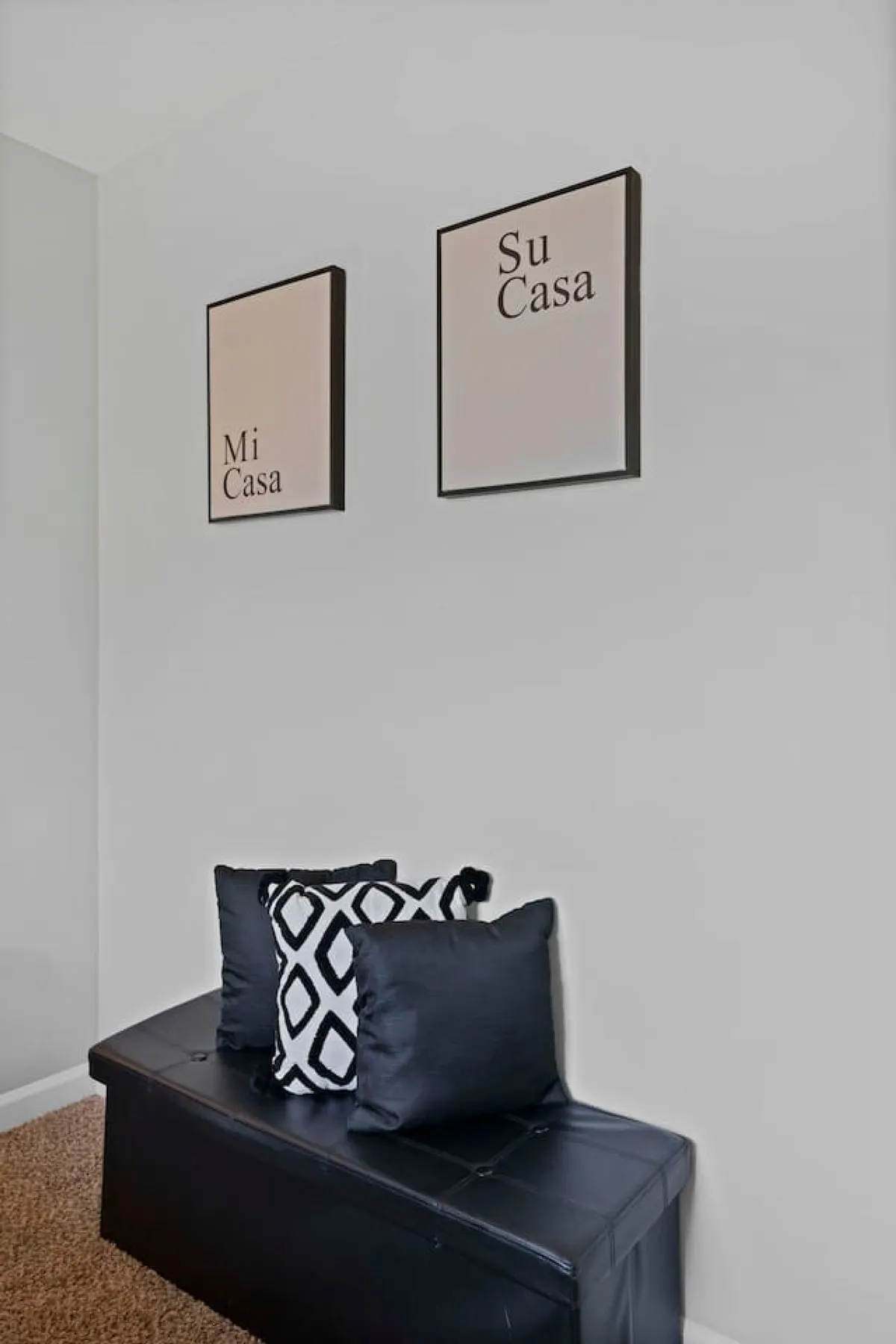 This image showcases a welcoming living space adorned with 'Mi Casa' and 'Su Casa' wall art, black and white decorative pillows, and a practical storage bench. The cozy seating area invites social interaction and relaxation, all set in a sunlit, friendly environment.