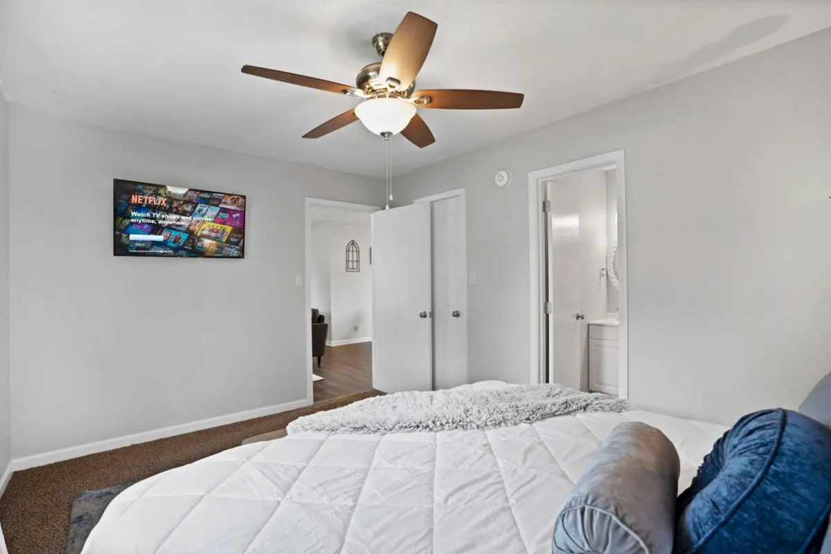 A cozy bedroom with white bedding, a television displaying Netflix, and a ceiling fan. The environment is comfortable and inviting, perfect for winding down after a night out.