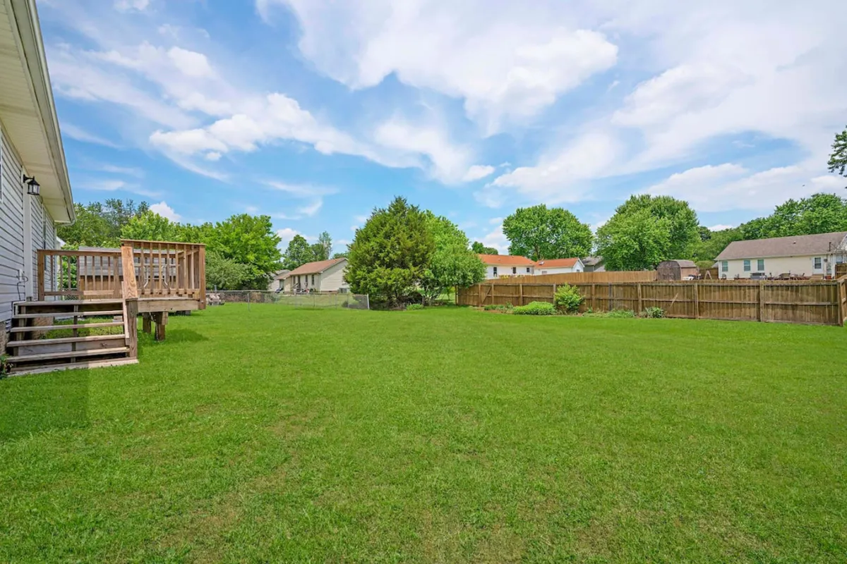 A wide view of a spacious backyard featuring a lush green lawn, a wooden deck, and mature trees framing the outdoor space. The natural sunlight highlights areas perfect for children's play, barbecues, or simply relaxing with a morning coffee amidst nature.