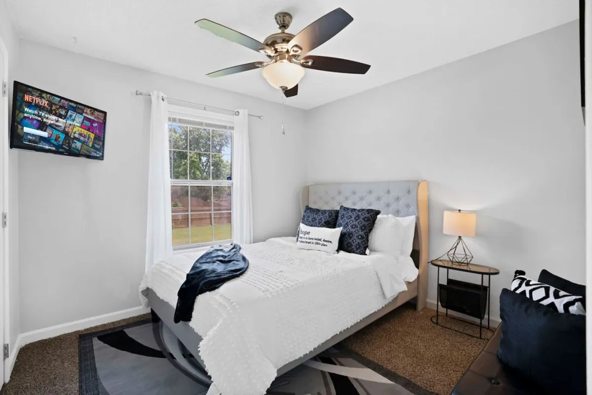 A cozy bedroom with a plush bed, soft lighting from a lamp, and a television, all framed by a window letting in natural light. The setting invites relaxation, whether for a movie night or a peaceful morning.