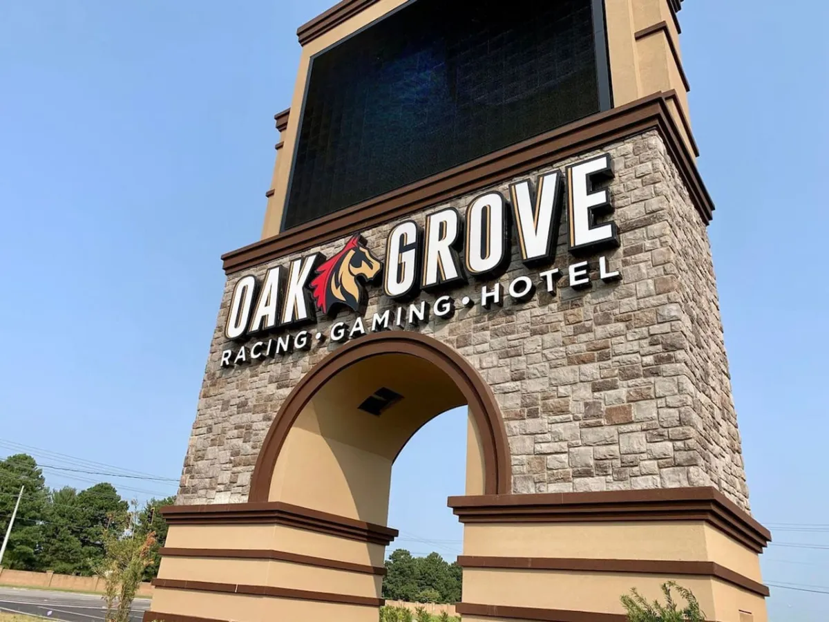 An exterior shot featuring the impressive 'OAK GROVE' sign beneath a stone archway with a digital display board, hinting at gaming, racing events, and hotel accommodations. The scene is lively and welcoming, evoking the excitement of a casino hotspot.