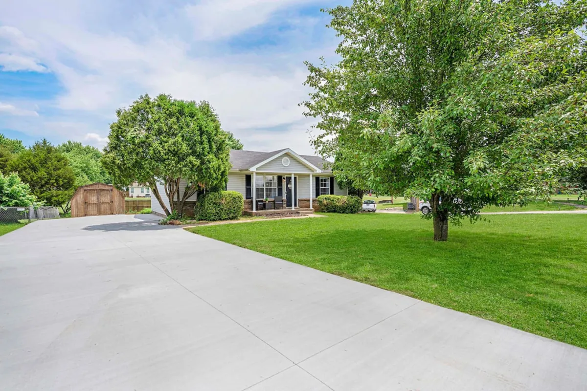 An inviting view of a home's front porch with a lush green lawn, mature trees, and a driveway. The scene conveys a sense of tranquility, safety, and the comforts of home, ideal for gatherings or relaxing after a busy day.