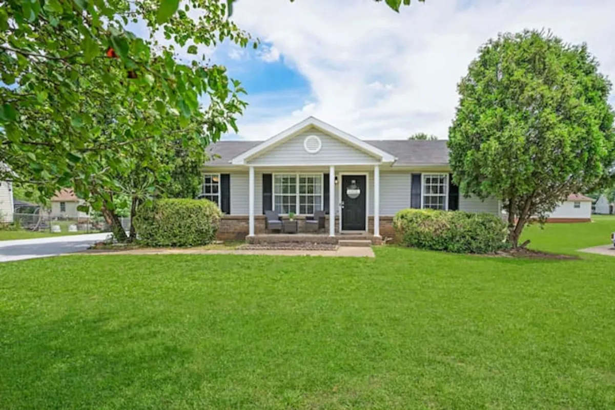 The image shows the charming exterior of a single-family house with a welcoming front porch, a lush green lawn, and mature trees creating a peaceful setting. The porch is perfect for relaxing with morning coffee, while the spacious yard invites children to play and families to gather outdoors.