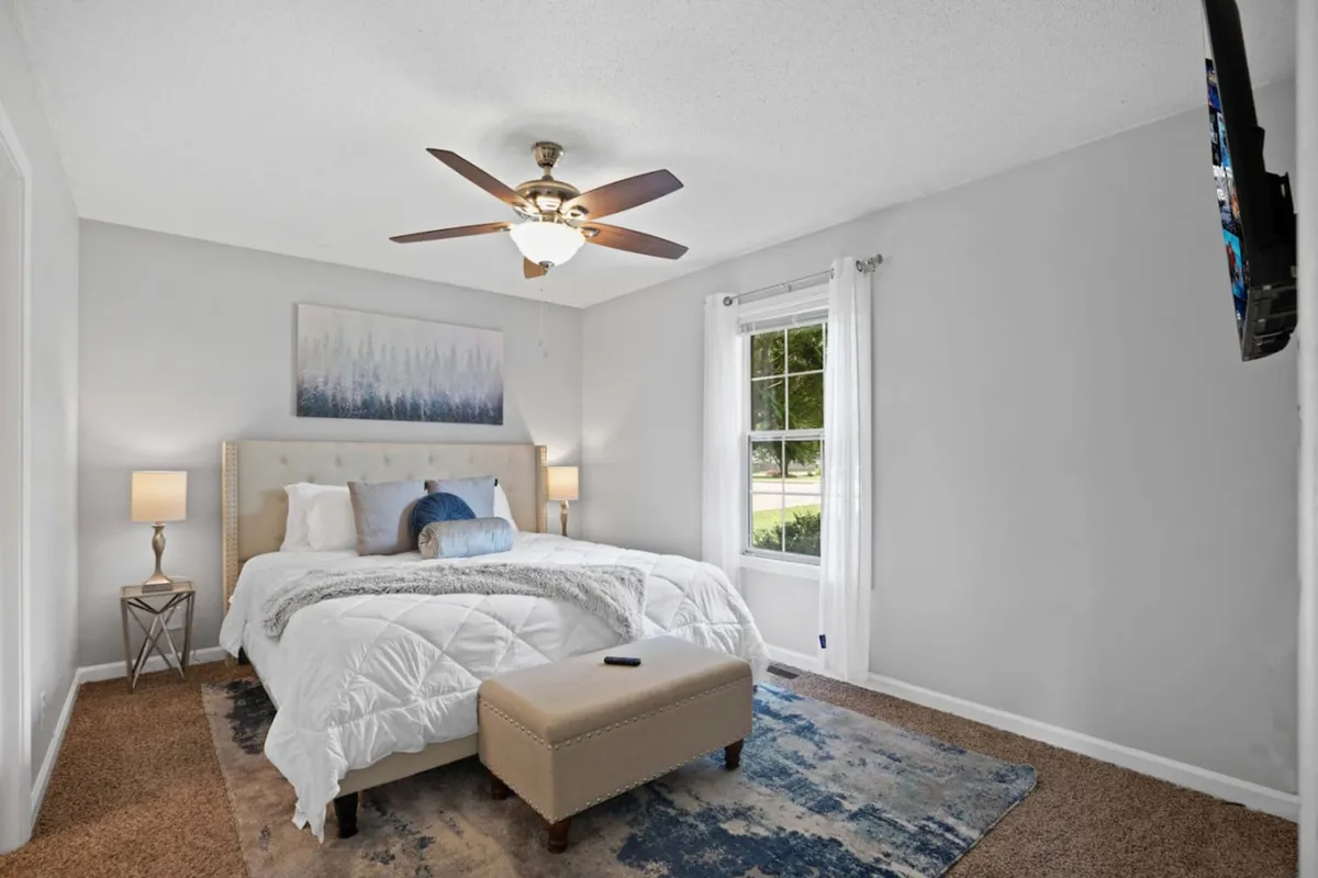 A tranquil bedroom scene with a cozy bed adorned with decorative pillows, artwork above the bed, and an ottoman at the foot. A window allows sunlight to brighten the space, and a nightstand with a lamp completes the cozy setup.